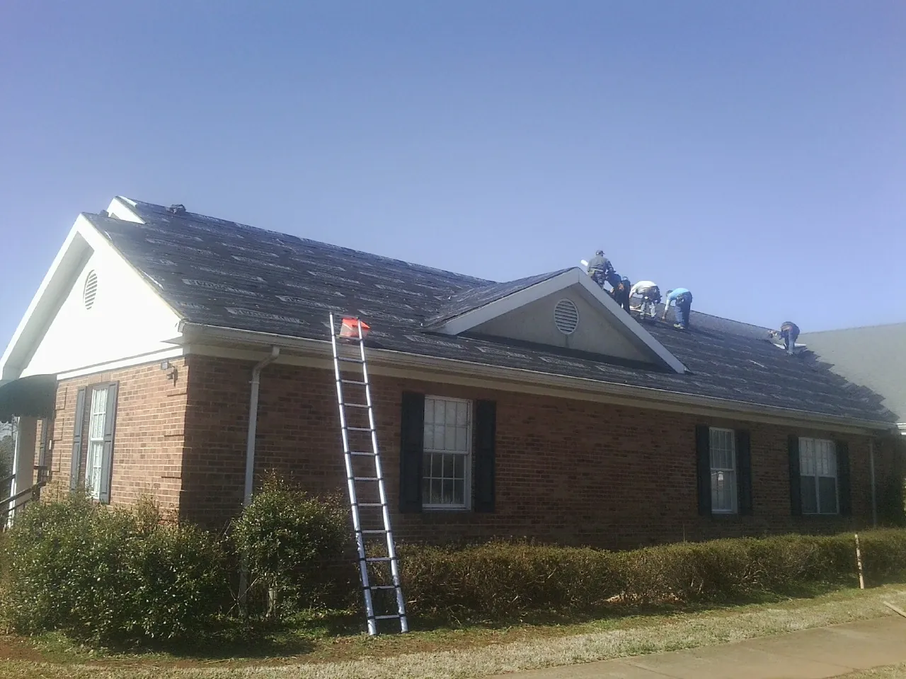 Crew tearing shingles off a brick ranch roof with a ladder set — Metal Roof Repair work by our crew in East Lynne