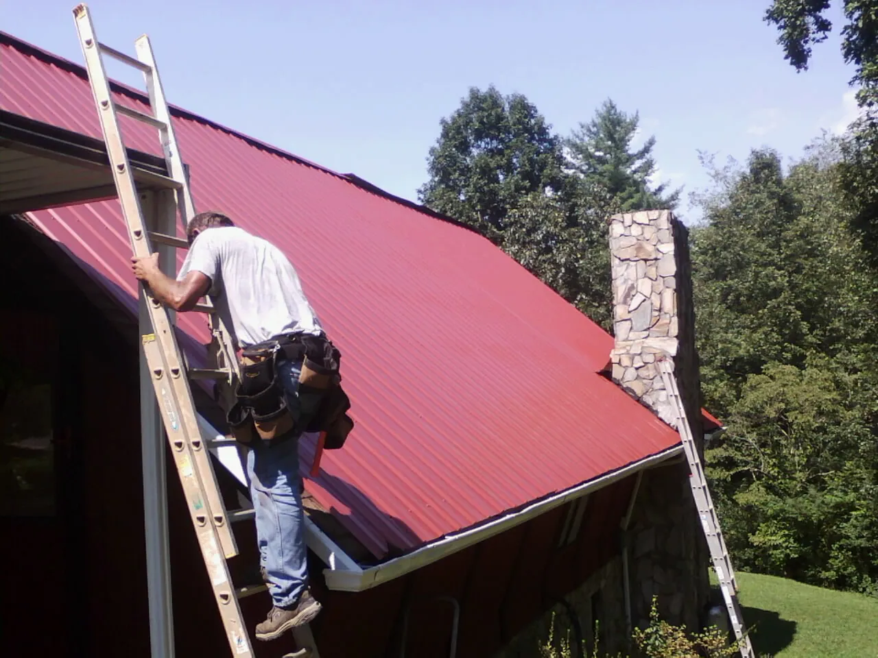 Roofer on a ladder beside a red metal roof and stone chimney — Soffit & Fascia Repair by Roofing Brothers in East Lynne